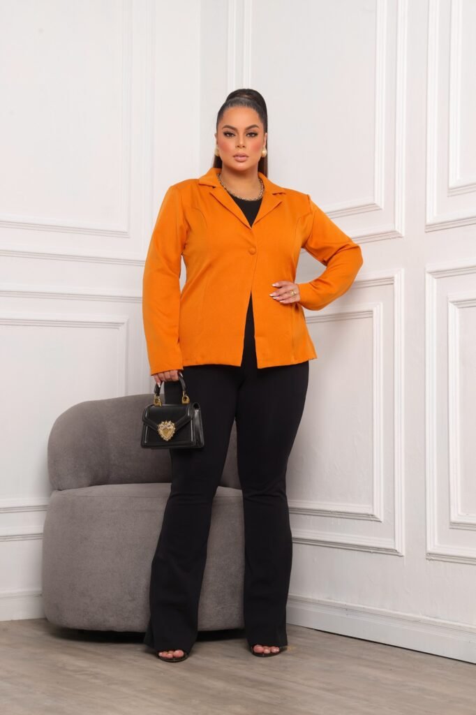 A fotografia mostra uma modelo plus, com uma calça comprida na cor preta e com um blazer na cor laranja, segurando uma clutch preta e cabelo preso.