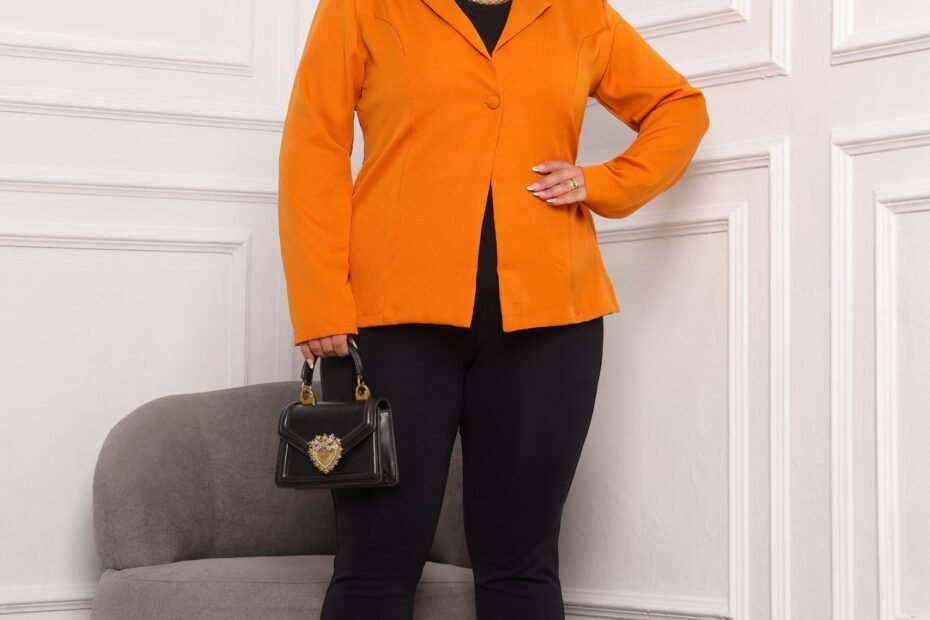 A fotografia mostra uma modelo plus, com uma cal&ccedil;a comprida na cor preta e com um blazer na cor laranja, segurando uma clutch preta e cabelo preso.